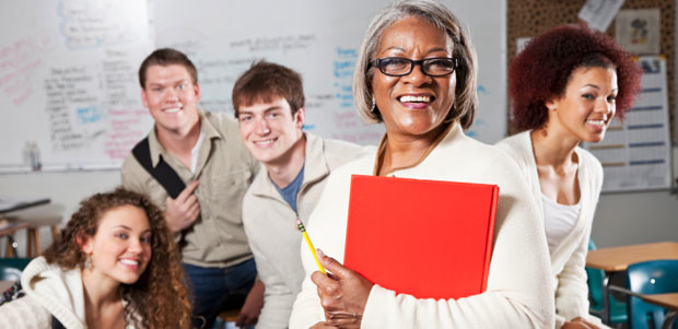 A smiling African American woman teacher stands in a classroom with happy high school students.